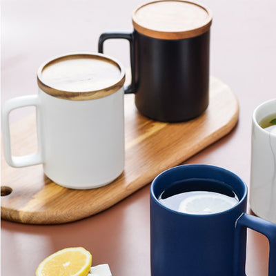 White, black and navy ceramic mug examples in a styled drinks scene, with wooden-edged lids shown on two mugs